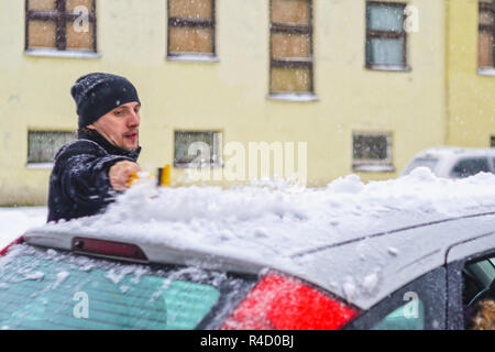 Giovane uomo in cappotto nero pulisce la sua vettura con il pennello di colore giallo durante la nevicata. Inverno condizioni atmosferiche avverse. Foto Stock