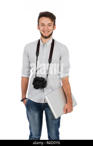 Il ragazzo del Nerd con fotocamera intorno al suo collo e computer sotto il suo braccio, è in piedi in studio e indossa abiti casual Foto Stock