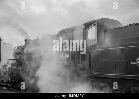 Bianco e nero, atmosfera, vista laterale da vicino della vecchia locomotiva a vapore del Regno Unito d'epoca in attesa della partenza, accanto alla piattaforma, che rilascia vapore. Foto Stock