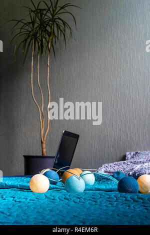 Grigio Laptot posa sul materasso con coperchio color turchese, alcuni cuscini e uno sfondo bianco - con un impianto in camera oltre ad esso e decorazione di Natale Luci Foto Stock