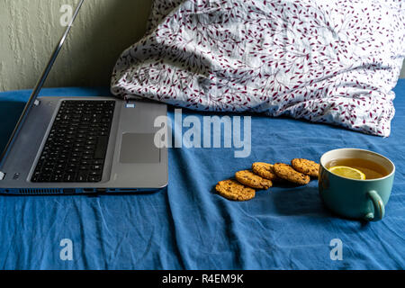 Grigio Laptot posa sul materasso con coperchio color turchese, alcuni cuscini e uno sfondo bianco - con tè e biscotti oltre ad esso Foto Stock