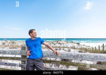 Destin, STATI UNITI D'AMERICA Miramar Beach City town village giorno in Florida Panhandle golfo del Messico di acqua oceanica, giovane uomo felice in maglietta blu appoggiata sulla recinzione di legno Foto Stock