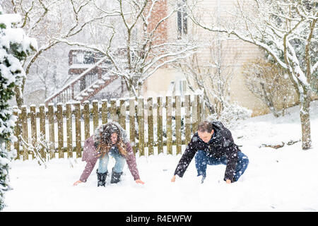 Giovane uomo, donna giocando, seduto, rendendo snowballs in inverno tempesta di neve, Storm, nevicava a casa, casa giardino cortile anteriore, nel cortile alberi coperti in Foto Stock