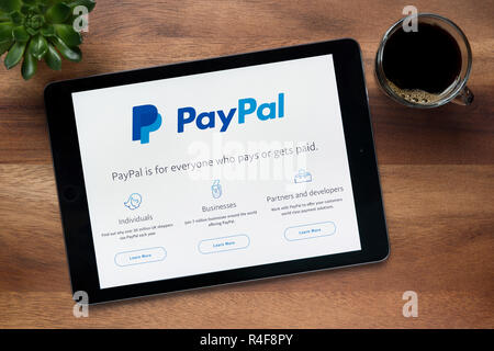 Il sito web di PayPal è visto su un tablet iPad, su di un tavolo di legno lungo con un caffè espresso e una pianta di casa (solo uso editoriale). Foto Stock