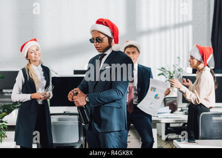 I giovani africani imprenditore americano in santa cappello e occhiali da sole di aprire una bottiglia di vino mentre festeggia il Natale con i colleghi in ufficio Foto Stock