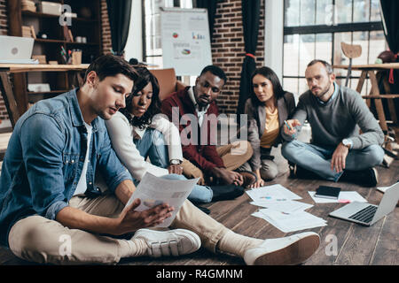 Grave gruppo multietnico di colworkers seduto sul pavimento e discutendo insieme un nuovo progetto in moderni loft office Foto Stock