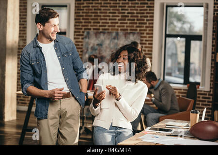 Multietnica paio di ridere e utilizza lo smartphone in ufficio loft con colleghi di lavoro dietro Foto Stock