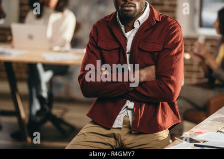 Vista ritagliata della african american casual uomo d affari seduto con le braccia incrociate e colleghi lavorando dietro in ufficio loft Foto Stock