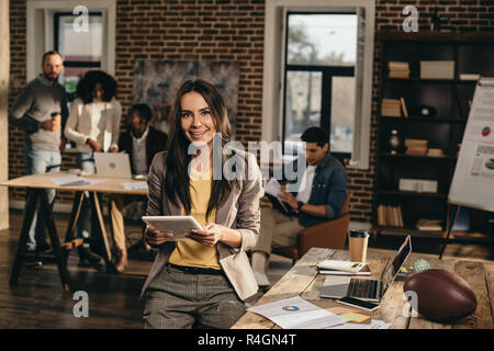 Sorridente casual imprenditrice azienda tablet in ufficio loft con colleghi lavorando dietro Foto Stock