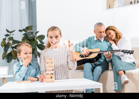 Sorella e fratello la riproduzione di blocchi di legno torre di gioco mentre i loro nonni giocare su chitarra dietro a casa Foto Stock