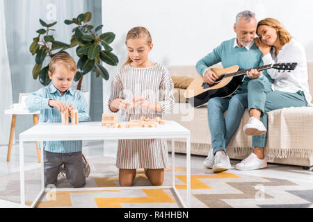 Messa a fuoco selettiva dei bambini che giocano con i blocchi di legno torre di gioco mentre i loro nonni giocare su chitarra dietro a casa Foto Stock