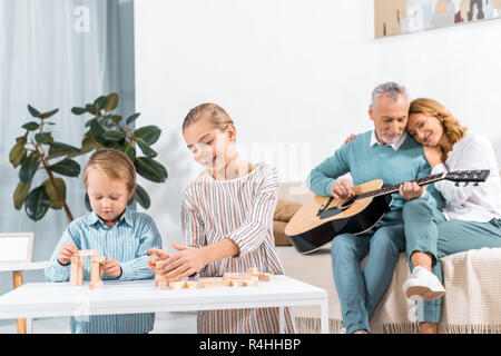 Felice sorella e fratello la riproduzione di blocchi di legno torre di gioco mentre i loro nonni giocare su chitarra dietro a casa Foto Stock