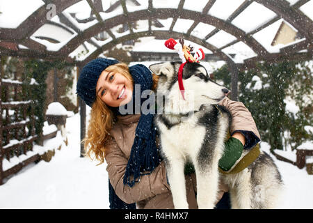 Attraente autentica donna caucasica abbracci funny Malamute Cane indossando santa cari natale palchi. Ricci femmina sorridente divertendosi con huskie cucciolo sul nuovo anno. Il Pet è regalo migliore per le vacanze. Foto Stock