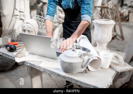 Scultore lavorare con computer portatile presso il posto di lavoro nel vecchio studio con sculture sullo sfondo. Immagine ritagliata con nessun volto Foto Stock