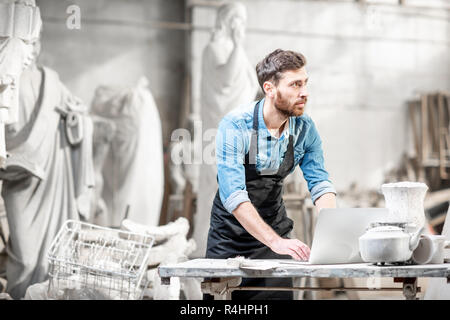 Scultore bello lavorare con computer portatile presso il posto di lavoro nel vecchio studio con sculture sullo sfondo Foto Stock