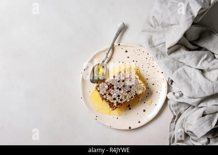 Miele biologico in favi con cucchiaio in spotted piastra ceramica sul panno di lino bianco su sfondo marmo. Laici piana, copia, spazio Foto Stock