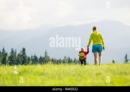 Felice madre con bambino camminando sul prato verde. Avventura Trekking con bambino in estate viaggio con la famiglia in montagna. Vacanze o weekend con attività Foto Stock