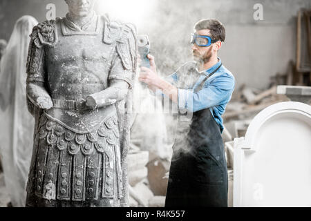 Scultore in protective workware macinazione pietra scultura con smerigliatrice electirc nel vecchio studio con polvere Foto Stock