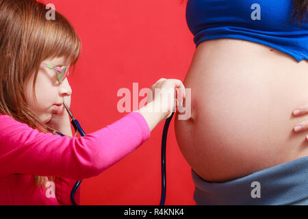 La gravidanza, il concetto di famiglia. Figlia utilizzando uno stetoscopio sulla sua madre gravida pancia per sentire i gemelli i toni del cuore Foto Stock