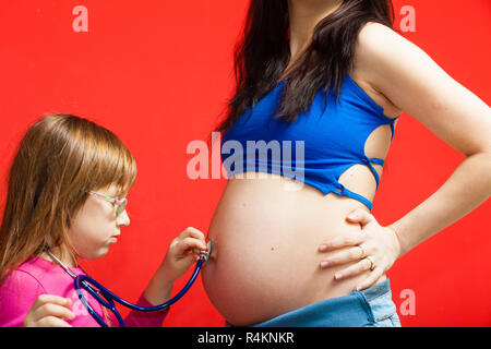 La gravidanza, il concetto di famiglia. Figlia utilizzando uno stetoscopio sulla sua madre gravida pancia per sentire i gemelli i toni del cuore Foto Stock