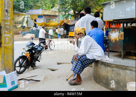 Il vecchio uomo indiano si siede presso la strada del mercato ed emissione di fumo di sigaretta. 11 febbraio 2018 Puttaparthi, India Foto Stock