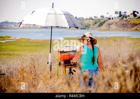 Sotto un ombrello sotto il sole, un artista femminile lavora sui plein air dipinto esterno che corrisponde alla sua vista panoramica di Newport Beach, CA, durante il tempo di concorso d'arte. (Foto di Spencer Grant) Foto Stock