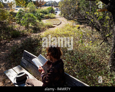 Milinial uomo riceve al di fuori di leggere in un parco pieno di piante autoctone, ancora circondato dal trambusto della vita urbana. Foto Stock