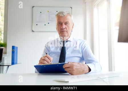 Ritratto di imprenditore senior seduto alla scrivania in ufficio e facendo alcuni documenti. Foto Stock