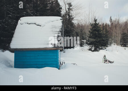 Una donna che si rilassa in una sedia adirondack di fronte alla sua piccola casa blu nella neve Foto Stock