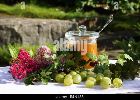 Atmosfera estiva con uva spina sul vassoio in legno amorevolmente decorate con fiori e vasetto di marmellata Foto Stock