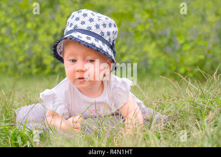 Ritratto di bimbo nel cappello. Baby ragazza seduta sul prato verde. Piccola principessa in abito bianco giacente su erba verde in giardino. La vacanza estiva b Foto Stock