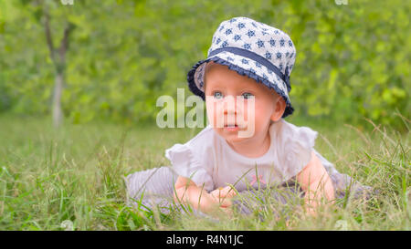 Baby toccando erba verde d'estate. Baby ragazza seduta sul prato verde. Piccola principessa in abito bianco giacente su erba verde in giardino. Ritratto Foto Stock