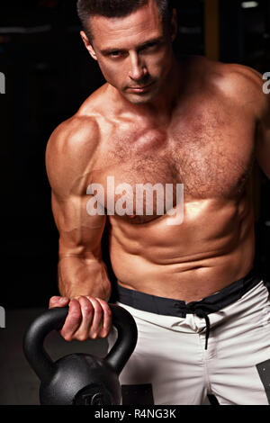 Forte arrabbiato athletic uomo facendo esercizi con kettlebells su ombra scura sullo sfondo. Closeup ritratto Foto Stock