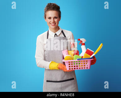 Ritratto di giovane sorridente scrubwoman nel grembiule con un cestello con detergenti organici e spazzole isolato su sfondo blu. Femmina. detergente per la pulizia Foto Stock