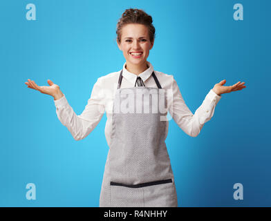 Sorridente scrubwoman giovani in grembiule presentare qualcosa su vuoto palm su sfondo blu. Scrubwoman in uniforme dimostra i servizi forniti da p Foto Stock