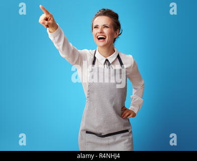 Sorridente scrubwoman giovani in grembiule puntando verso qualcosa su sfondo blu. Scrubwoman mostra sulle realizzazioni del servizio di pulizia nel lavoro wi Foto Stock