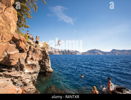 Giovani in attesa su di uno sperone di roccia e nuotare o saltare nel blu profondo delle acque di un lago di montagna, di Crater Lake, Oregon, Stati Uniti d'America Foto Stock