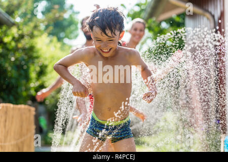 Ragazzo il raffreddamento con tubo flessibile da giardino, famiglia in background Foto Stock