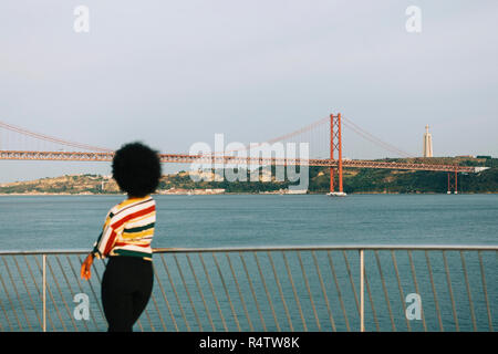 Giovane donna in piedi al Waterfront, guardando a 25 de Abril Bridge e Cristo Rei statua, Lisbona Foto Stock