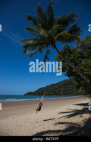 Carefree donna basculante in swing corda sotto la struttura Palm Tree sulla spiaggia tropicale, Port Douglas, Queensland Foto Stock
