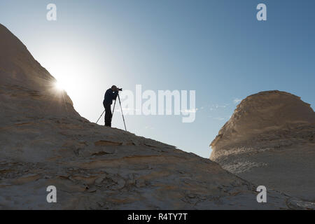 Silhouette di un fotografo di paesaggio riprese con un treppiede a Rising Sun nel suo retro in un epico paesaggio presso l'Egiziano deserto bianco Foto Stock