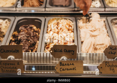 Fresco artigianale gelato il gelato in vendita in contenitori di metallo a mano con un cucchiaio di metallo come ottenere una porzione sullo sfondo. Foto Stock