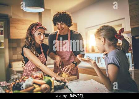 Genitori felici e la loro figlia cucina insieme in cucina mentre la bambina facendo il suo dovere sul tavolo della cucina. Foto Stock