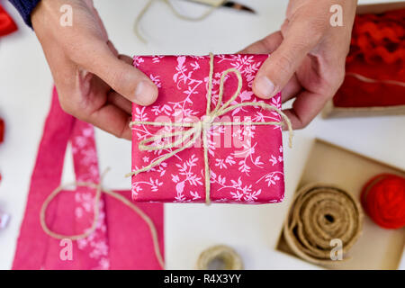 Elevato angolo di visione di un giovane uomo caucasico con un bel regalo in mano, avvolto in carta rosa e legato con un rustico di stringa, su un tavolo bianco pieno di Foto Stock