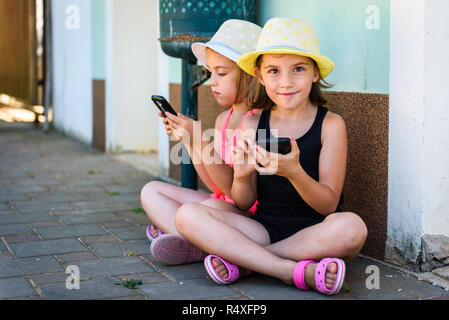 Gemelli identici utilizza lo smartphone per i giochi o internet per le vacanze. Bambine e bambini stanno giocando con lo smartphone, seduto sul pavimento lastricato in Foto Stock