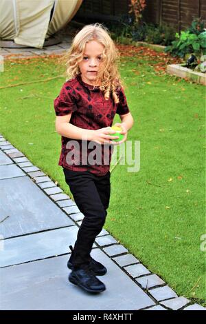 Un giovane, caucasica, capelli lunghi boy gioca con una palla in un giardino Foto Stock