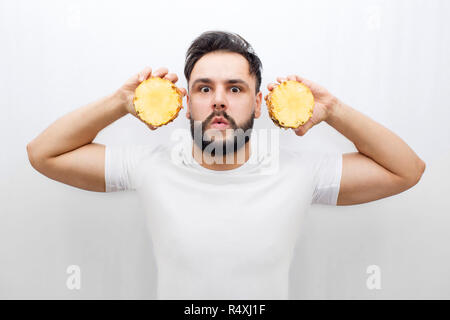Realmente sorpreso giovane uomo stand e guardare sulla fotocamera. Egli coveres orecchie con due halfs di uno ananas. Il ragazzo è spaventato e amased. Uomo in distratti. Isolato su sfondo bianco. Foto Stock