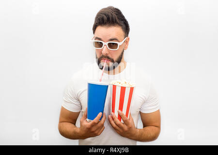 Giovane uomo si alza e guarda verso il basso. Indossa gli occhiali del filmato. Guy contiene carta vetro con popcorn e cola. Bevande egli attraverso la paglia e guardare sulla fotocamera. Isolato su sfondo bianco. Foto Stock