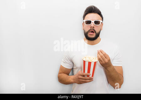 Entusiasti e stupiti giovane uomo si alza e guarda avanti sulla fotocamera. Indossa gli occhiali del filmato. Guy detiene il secchio di popcorn. La sua bocca è aperto. Isolato su sfondo bianco. Foto Stock