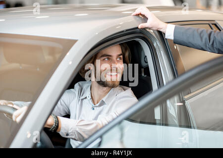 Giovane uomo cercando una nuova vettura seduto sul sedile lato guidatore a parlare con l'addetto alle vendite in showroom Foto Stock